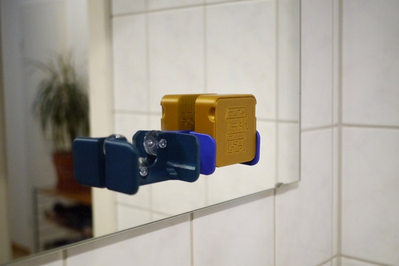 May include: Two gold-colored boxes with a design of a building on the front are attached to a blue and white wall mount. The wall mount is attached to a mirror.