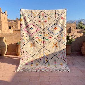 Puede incluir: Alfombra crema tejida a mano con un patrón geométrico de diamantes. El diseño presenta contornos negros y acentos coloridos en rosa, azul, amarillo y verde. La alfombra tiene un borde con flecos y se exhibe al aire libre.