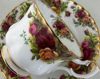 May include: White teacup and saucer set with a floral rose pattern in shades of red, yellow, and pink. The edges of the cup and saucer are trimmed with gold. The teacup has a curved handle.