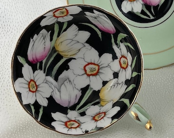 May include: A vintage teacup and saucer set. The teacup features a black interior with white floral designs and a gold rim. The saucer is light green with a black floral center and gold trim. The handle is light green with gold accents.