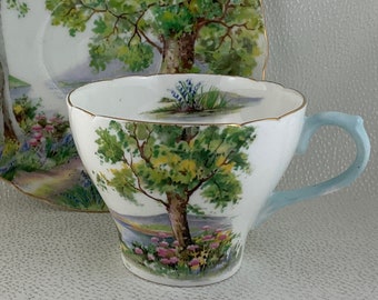 May include: A white teacup and saucer set with a landscape design. The cup has a light blue handle and gold trim. The saucer and cup feature a scenic view of trees, water, and flowers. The set is made of porcelain.