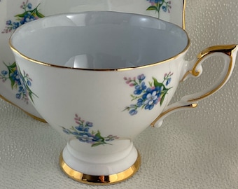 Könnte beinhalten: Weißes Tee-Set mit einer Tasse und Untertasse, verziert mit blauen und rosa Blumenmustern. Die Tasse hat einen goldenen Henkel und Fuß, die Untertasse einen gewellten Rand und Goldrand.