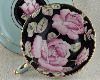 May include: A vintage teacup and saucer set. The teacup features a black interior adorned with pink roses and green leaves. The saucer is light blue with a gold rim and a central black circle with a pink rose. The handle is light blue with gold accents.