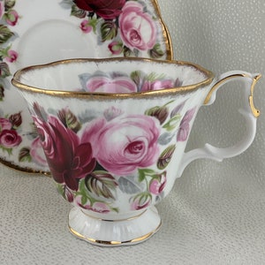 Puede incluir: Juego de taza y platillo blancos con un patrón de rosas en tonos rosa y burdeos. El conjunto tiene ribetes dorados en los bordes y el asa. El platillo tiene un borde festoneado.