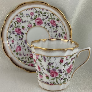 May include: A white teacup and saucer set with a floral pattern of pink roses and purple accents. The edges are trimmed with gold. The cup has a unique, faceted shape.