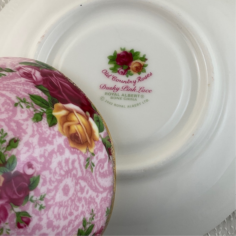 May include: A close-up of a Royal Albert bone china teacup and saucer set. The teacup features a pink lace pattern with roses in shades of pink and yellow. The saucer is white with the text "Old Country Roses Dusky Pink Lace".
