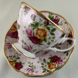 May include: A vintage teacup and saucer set. The cup and saucer are decorated with a pink and white pattern and a floral design featuring yellow, red, and pink roses. The edges are trimmed with gold.