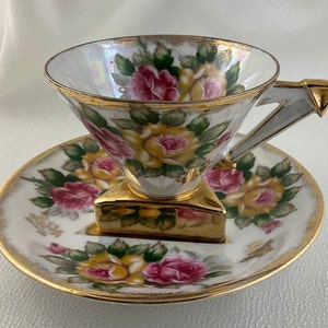 Puede incluir: Un juego de taza y platillo de té vintage. La taza y el platillo de porcelana blanca están adornados con patrones florales de rosas amarillas y rosas, acentuados con ribetes dorados. La taza tiene un asa geométrica única y se apoya en una base dorada.