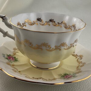 May include: A vintage teacup and saucer set. The teacup and saucer are white with a yellow gradient and gold trim. The teacup has a curved handle and the saucer has a scalloped edge. Pink roses are painted on the saucer.