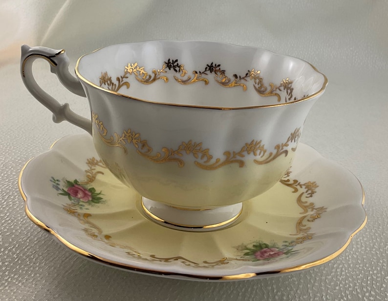 May include: A vintage teacup and saucer set. The teacup and saucer are white with a yellow gradient and gold trim. The teacup has a curved handle and the saucer has a scalloped edge. Pink roses are painted on the saucer.