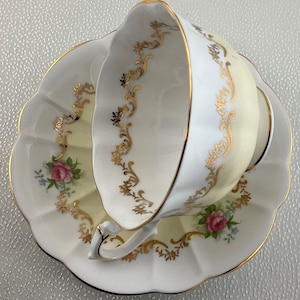 May include: A white teacup and saucer set with a scalloped edge. The cup and saucer have a pale yellow interior and are decorated with gold filigree and pink roses. The rim is trimmed with gold.