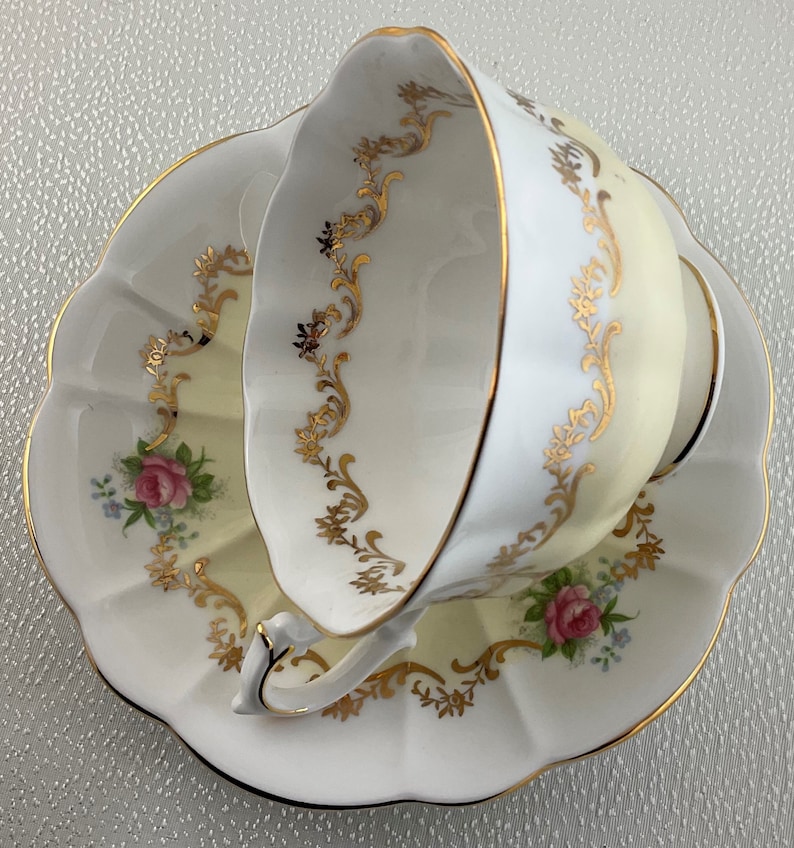 May include: A white teacup and saucer set with a scalloped edge. The cup and saucer have a pale yellow interior and are decorated with gold filigree and pink roses. The rim is trimmed with gold.