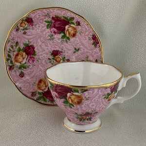 May include: A pink teacup and saucer set with a floral rose pattern. The cup and saucer have gold trim. The teacup has a white interior and a pedestal base. The saucer is round with a scalloped edge.