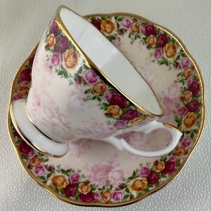 May include: A porcelain teacup and saucer set. The cup and saucer are decorated with a floral pattern of pink, yellow, and burgundy roses. The edges of the cup and saucer are trimmed with gold. The cup is tilted, revealing its white interior.