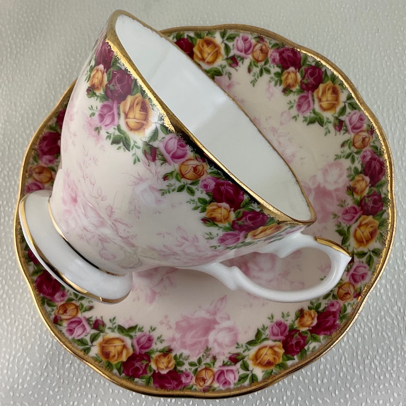 May include: A porcelain teacup and saucer set. The cup and saucer are decorated with a floral pattern of pink, yellow, and burgundy roses. The edges of the cup and saucer are trimmed with gold. The cup is tilted, revealing its white interior.