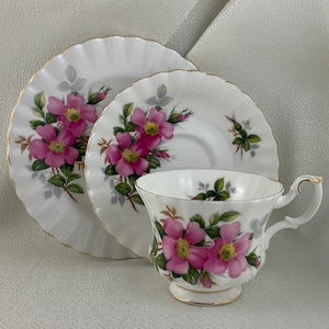 Puede incluir: Juego de té blanco con taza, platillo y plato. El juego presenta un diseño floral con flores rosas, hojas verdes y ribete dorado. El platillo y el plato tienen un borde acanalado.