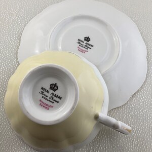 May include: A Royal Albert Bone China teacup and saucer set. The teacup is pale yellow with a scalloped edge, and the saucer is white with a matching scalloped edge. The set is from the Invitation Series, with the Royal Albert logo visible.