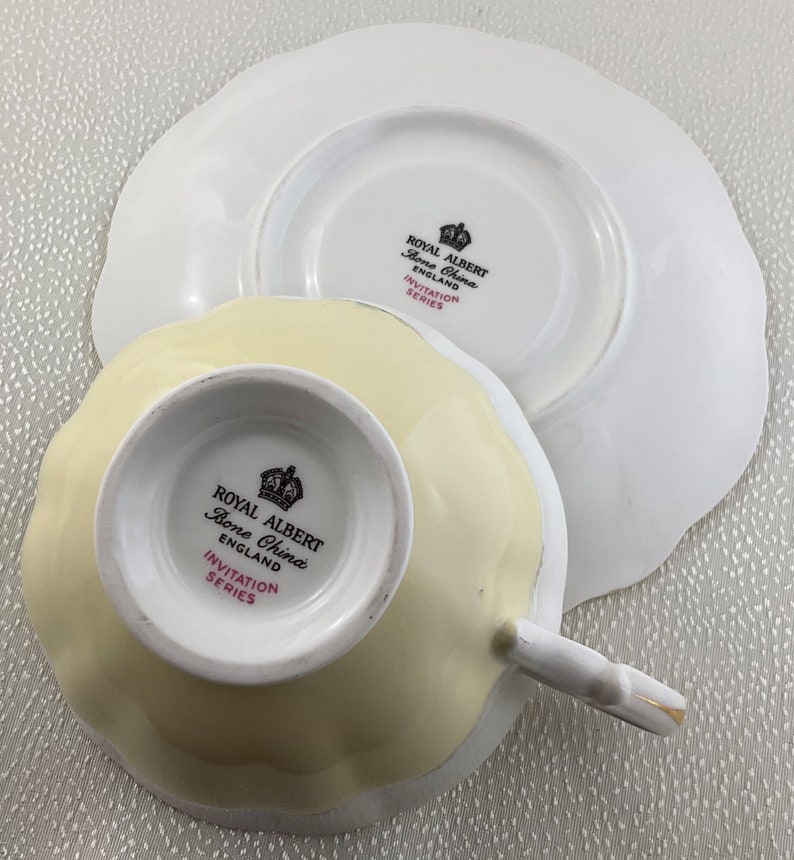 May include: A Royal Albert Bone China teacup and saucer set. The teacup is pale yellow with a scalloped edge, and the saucer is white with a matching scalloped edge. The set is from the Invitation Series, with the Royal Albert logo visible.