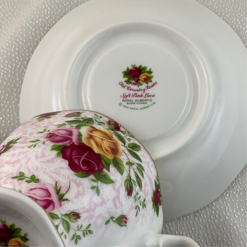 May include: A white teacup and saucer set with a floral pattern of pink, red, and yellow roses. The teacup has a curved handle and the saucer is round. The set is marked "Old Country Roses Soft Pink Lace" and "Royal Albert Bone China."