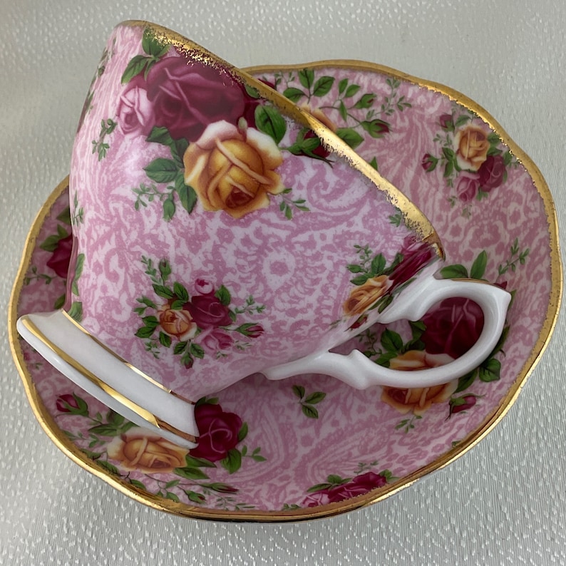 May include: A pink teacup and saucer set with a floral rose pattern. The cup and saucer have gold trim and a white handle. The roses are in shades of yellow, red, and pink.