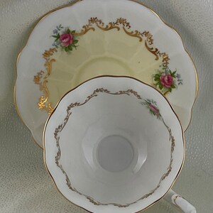 May include: A vintage teacup and saucer set. The saucer has a pale yellow center and is decorated with pink roses, blue flowers, and gold trim. The teacup is white with gold trim and a scalloped edge. A classic tea set.
