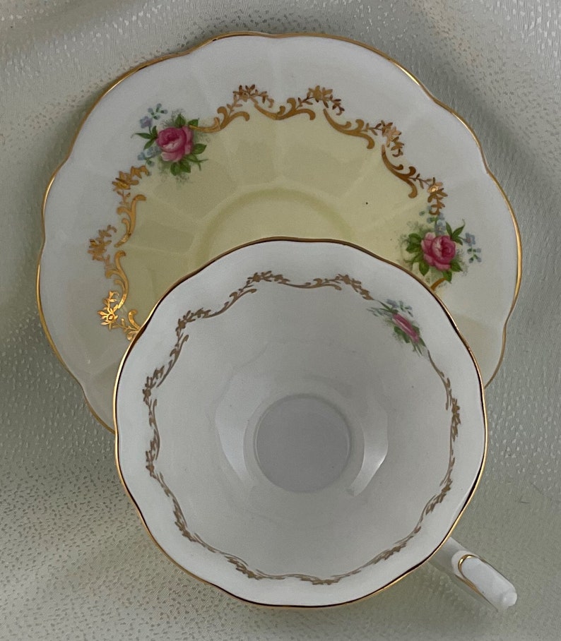May include: A vintage teacup and saucer set. The saucer has a pale yellow center and is decorated with pink roses, blue flowers, and gold trim. The teacup is white with gold trim and a scalloped edge. A classic tea set.