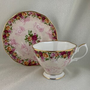 May include: A white teacup and saucer set with a floral rose pattern in pink, yellow, and burgundy. The cup and saucer have gold trim. The saucer has a scalloped edge.