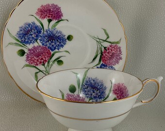 May include: White teacup and saucer set with a floral design. The cup and saucer feature a gold rim and a pattern of blue and pink flowers with green stems and buds. The teacup has a pedestal base.