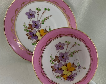 May include: A vintage teacup and saucer set. The set has a pink rim with a gold scalloped edge. The interior of the cup and saucer are white with a floral design of purple, yellow, and brown flowers.