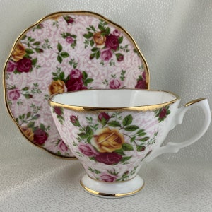 May include: A white teacup and saucer set with a floral rose pattern in shades of pink, yellow, and burgundy. The edges are trimmed with gold. The teacup has a pedestal base and a curved handle.