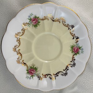 May include: A white and yellow scalloped saucer with a gold rim. The saucer features pink roses and blue flowers with gold and black decorative accents. The centre of the saucer is a pale yellow colour.