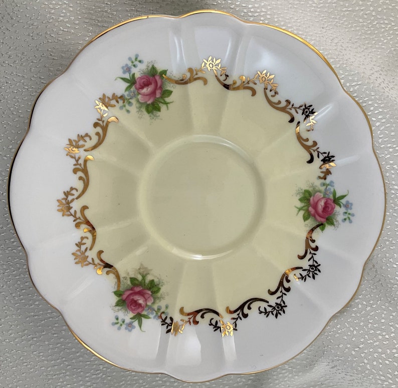 May include: A white and yellow scalloped saucer with a gold rim. The saucer features pink roses and blue flowers with gold and black decorative accents. The centre of the saucer is a pale yellow colour.