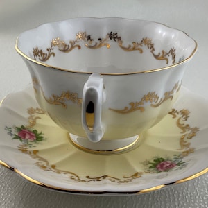 May include: A vintage teacup and saucer set. The cup and saucer are yellow and white with gold trim and floral details. The cup has a curved handle and a scalloped rim. The saucer has a matching scalloped edge.