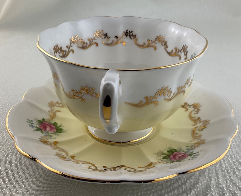 May include: A vintage teacup and saucer set. The cup and saucer are yellow and white with gold trim and floral details. The cup has a curved handle and a scalloped rim. The saucer has a matching scalloped edge.
