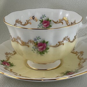 May include: A vintage teacup and saucer set. The cup and saucer are white with a pale yellow base, decorated with pink roses, blue flowers, and gold detailing. The rim and handle are edged with gold.