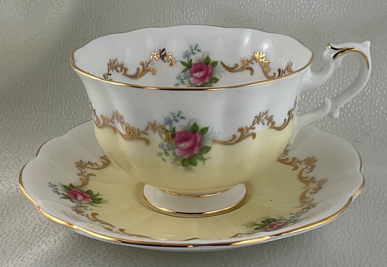 May include: A vintage teacup and saucer set. The cup and saucer are white with a pale yellow base, decorated with pink roses, blue flowers, and gold detailing. The rim and handle are edged with gold.