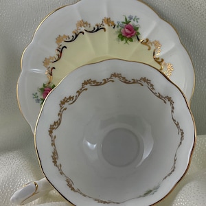 May include: A white teacup and saucer set with gold trim and floral details. The cup and saucer have a scalloped edge. The saucer features a pink rose and blue flower bouquet. The cup has a gold filigree design around the rim.