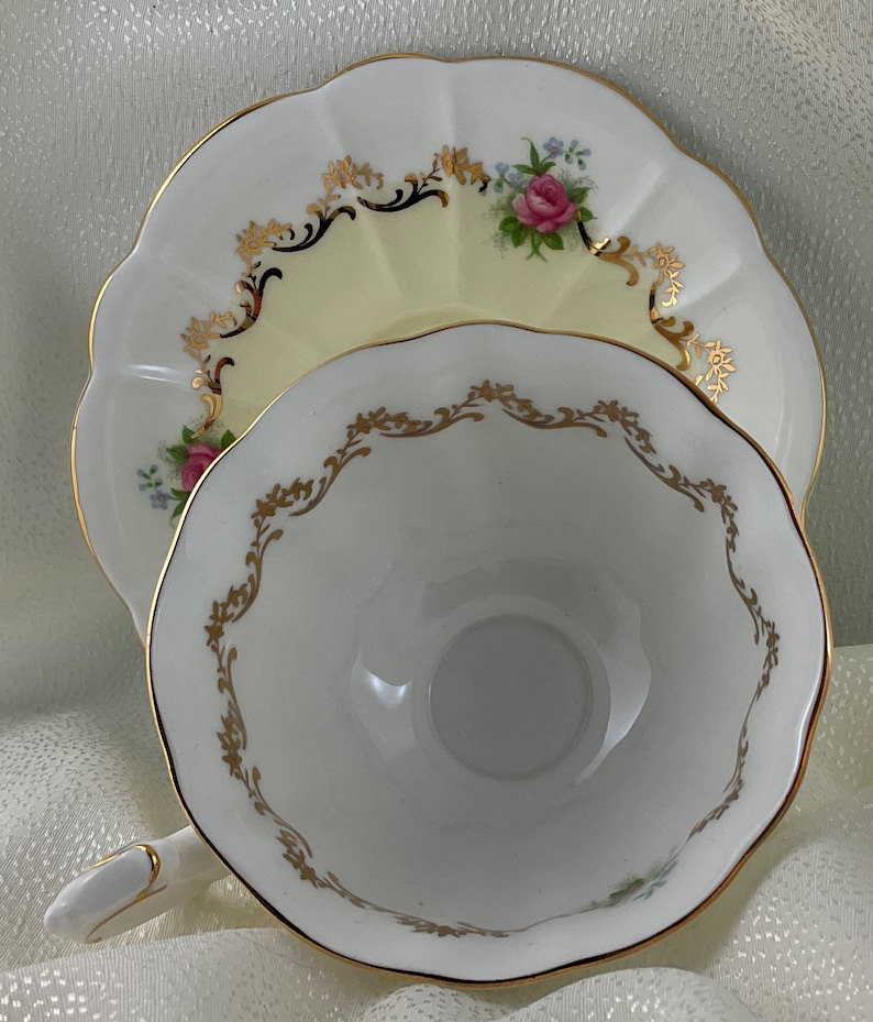 May include: A white teacup and saucer set with gold trim and floral details. The cup and saucer have a scalloped edge. The saucer features a pink rose and blue flower bouquet. The cup has a gold filigree design around the rim.