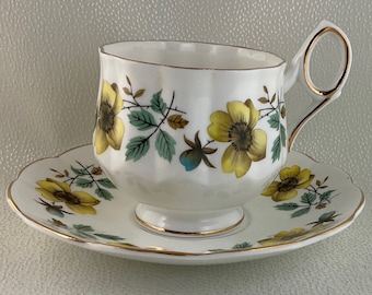 May include: White teacup and saucer set with a floral design. The cup and saucer feature yellow flowers, green leaves, and gold trim. The cup has a unique handle design.
