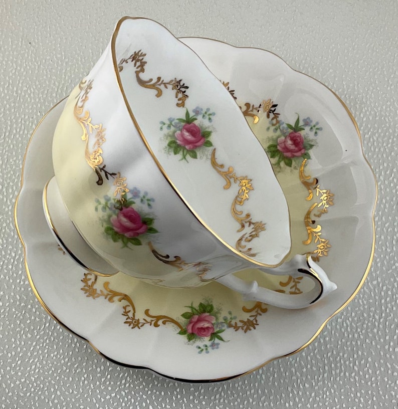 May include: A white teacup and saucer set with a scalloped edge. The set features pink roses and gold filigree accents. The teacup is tilted, revealing the interior. A classic design for tea time.