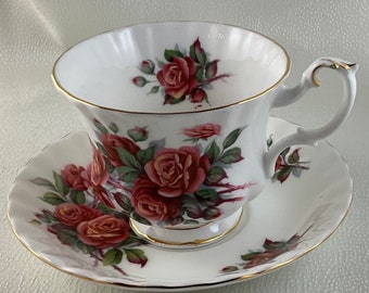 Puede incluir: Juego de taza y platillo blancos con un diseño floral de rosas rojas y hojas verdes. La taza y el platillo tienen ribetes dorados. El asa de la taza es curvada.