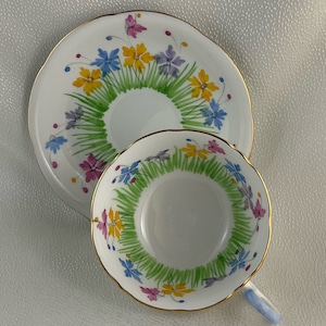 Op de afbeelding: Witte theekop en schotel set met een bloemenontwerp. De set heeft een ring van groen gras met gele, blauwe en paarse bloemen. De kop heeft een lichtblauw handvat en gouden rand. De schotel heeft een geschulpte rand.
