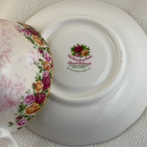 May include: A white teacup and saucer set with a floral design. The cup and saucer are decorated with pink, yellow, and red roses. The saucer has the text "Old Country Roses Peach Damask" and "Royal Albert Bone China".