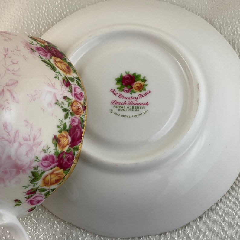 May include: A white teacup and saucer set with a floral design. The cup and saucer are decorated with pink, yellow, and red roses. The saucer has the text "Old Country Roses Peach Damask" and "Royal Albert Bone China".