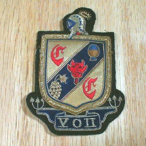 May include: Embroidered patch featuring a shield design with a devil, grapes, and other symbols. The patch has gold, red, blue, and black accents, with the text "VOH" at the bottom. A knight's helmet is at the top.