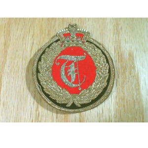 May include: A circular emblem with a red center featuring a silver letter "T" and an anchor. The emblem is surrounded by a gold wreath and a black border, topped with a gold crown. The background is a light wood grain.