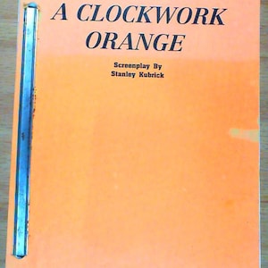 May include: An orange screenplay titled "A Clockwork Orange" by Stanley Kubrick. The title is in large, bold, black font. The screenplay is bound with a silver metal strip along the left edge.
