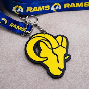 May include: A yellow and black keychain with the Los Angeles Rams logo, a ram's head. The keychain is attached to a blue lanyard with the word "RAMS" printed on it. The lanyard has a repeating pattern of the team logo.