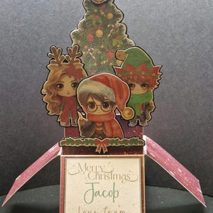 May include: A pop-up Christmas card featuring a Christmas tree with a golden snitch on top, a Harry Potter character, an elf, and a reindeer. The card is open and says "Merry Christmas Jacob Love from Nanna xx".