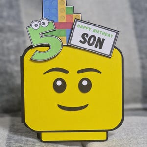 May include: A yellow birthday card in the shape of a Lego head, featuring a green number 5 and a sign reading "Happy Birthday Son." The card is adorned with colourful Lego bricks.